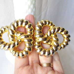 May include: Two gold-toned, intertwined knot-shaped brooches. The brooches have a textured, rope-like design and are held in a hand. The background is a soft, white fabric.