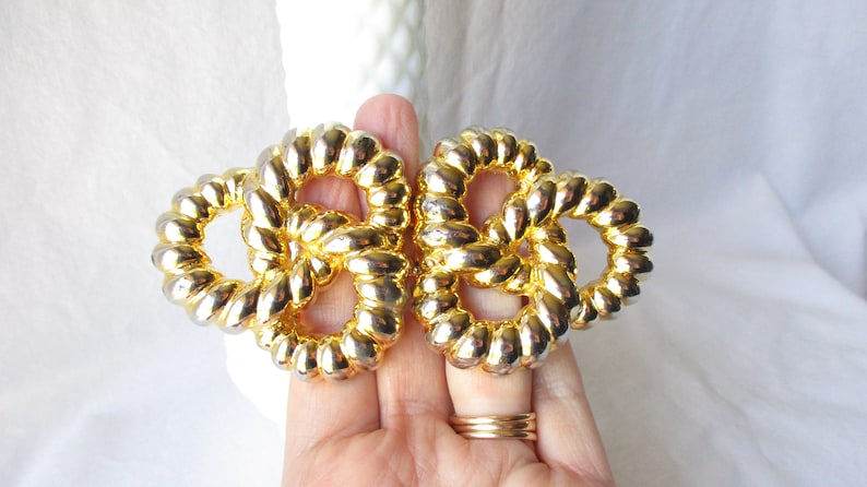May include: Two gold-toned, intertwined knot-shaped brooches. The brooches have a textured, rope-like design and are held in a hand. The background is a soft, white fabric.