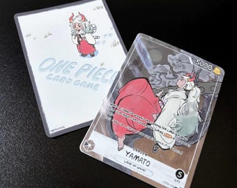 Puede incluir: Dos cartas coleccionables de One Piece con el personaje Yamato. Una carta muestra a Yamato en estilo chibi con el texto One Piece Card Game. La otra carta muestra a Yamato en una pose dinámica con el texto Leader Yamato, 5000 power y 5 life.