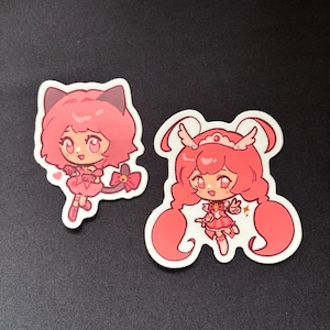 May include: Two die-cut vinyl stickers featuring chibi-style anime characters. The left sticker shows a pink-haired character with cat ears and a tail. The right sticker depicts a pink-haired character with a crown, long pigtails, and wings.