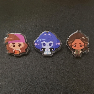 May include: Three clear acrylic pins featuring cartoon characters. One pin has a character with brown hair and a pink hat. Another has a blue-haired character. The third pin has a character with brown hair and a green outfit.