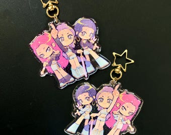 May include: Two clear acrylic keychains featuring a cartoon illustration of three stylized figures with pink and purple hair, wearing matching outfits. Each keychain has a gold star charm and a gold ring.