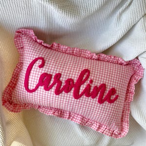 May include: A rectangular pink and white gingham pillow with the name "Caroline" embroidered in a vibrant pink script. The pillow has a ruffled edge and is set against a white textured background.