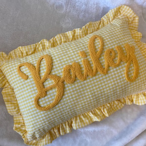 Könnte beinhalten: Ein rechteckiges Kissen mit gelb-weißem Vichy-Muster. Das Wort "Bailey" ist in gelber Schrift über die Mitte gestickt. Das Kissen hat einen gerüschten gelben Rand.