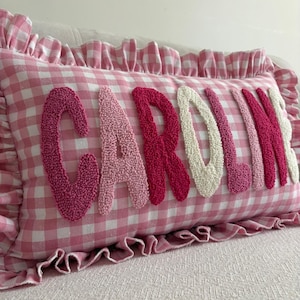 Könnte beinhalten: Ein rechteckiges Zierkissen mit rosa-weißem Vichy-Karo-Muster und einem gerüschten Rand. Der Name CAROLINE ist in erhabener, strukturierter Punch-Needle-Stickerei in abwechselnden Farbtönen von Dunkelrosa, Hellrosa und Creme aufgestickt.