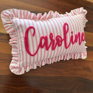 May include: A rectangular pillow with pink and white stripes and the name "Caroline" embroidered in a cursive font. The pillow has a ruffled edge in a matching pink and white striped fabric.