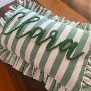 May include: A rectangular throw pillow featuring a sage green and white vertical striped pattern. The pillow is accented with a ruffled border along the edges and displays the name Clara in raised, textured dark green cursive embroidery.