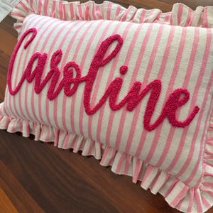 May include: A rectangular pillow with pink and white stripes and a ruffled edge. The name "Caroline" is embroidered in a cursive font in a vibrant pink color across the center of the pillow. The pillow is on a wooden surface.