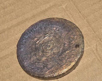 May include: A circular, dark bronze-toned metal pendant with a textured, uneven surface featuring a central relief design. The object has a small, circular hole near the right edge for attachment, with a weathered, metallic finish.