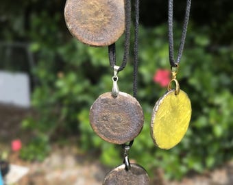 May include: Four circular pendants hang from black cords. The top pendant is textured brown, the middle left is dark brown, the middle right is bright yellow, and the bottom pendant is a metallic gray. Each piece features a metal bail.