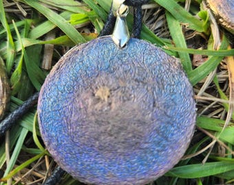 May include: A round, blue and purple pendant with a silver bail. The pendant is made of resin and has a textured surface. It is hanging from a black cord.