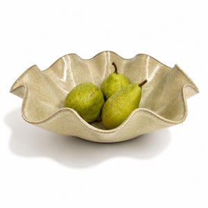 May include: A beige ceramic bowl with a wavy, ruffled edge containing three green pears. The bowl has a speckled, textured finish and a glossy glaze. The pears are smooth with brown stems, positioned inside the centre of the wide, shallow vessel.