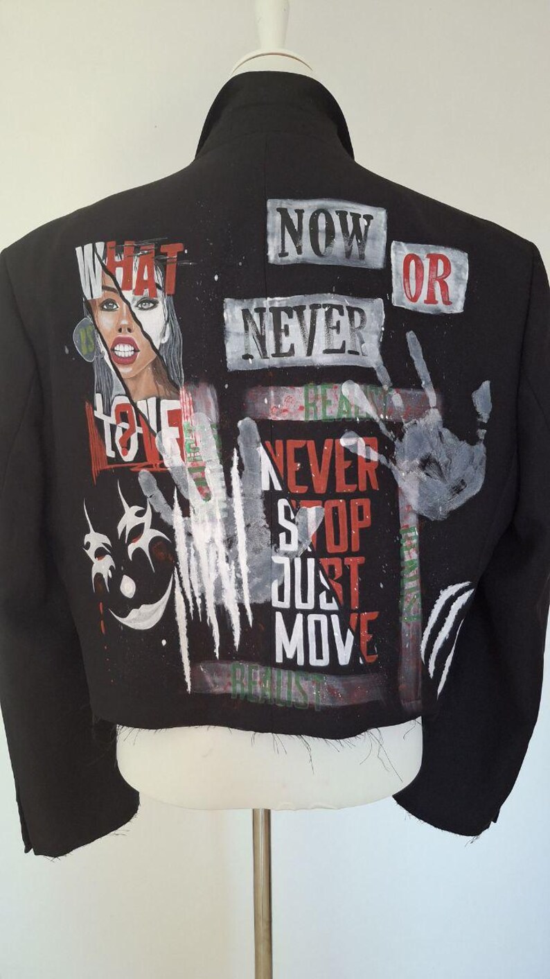 K&ouml;nnte beinhalten: Schwarze, kurze Jacke mit Graffiti-Design. Sie zeigt die Worte "NOW OR NEVER", "NEVER STOP JUST MOVE" und eine Grafik eines Frauengesichts. Die Jacke hat einen klassischen Kragen und wird auf einer Schaufenquin pr&auml;sentiert.