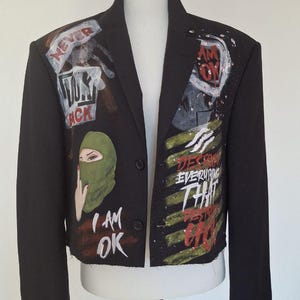 K&ouml;nnte beinhalten: Schwarzer Blazer mit handgemalten Kunstwerken. Die Jacke zeigt eine gr&uuml;ne Sturmhaube, die Worte "I AM OK" und andere Phrasen. Das Kunstwerk ist in Rot, Wei&szlig; und Gr&uuml;n gehalten. Der Blazer wird auf einer wei&szlig;en Schaufensterpuppe pr&auml;sentiert.