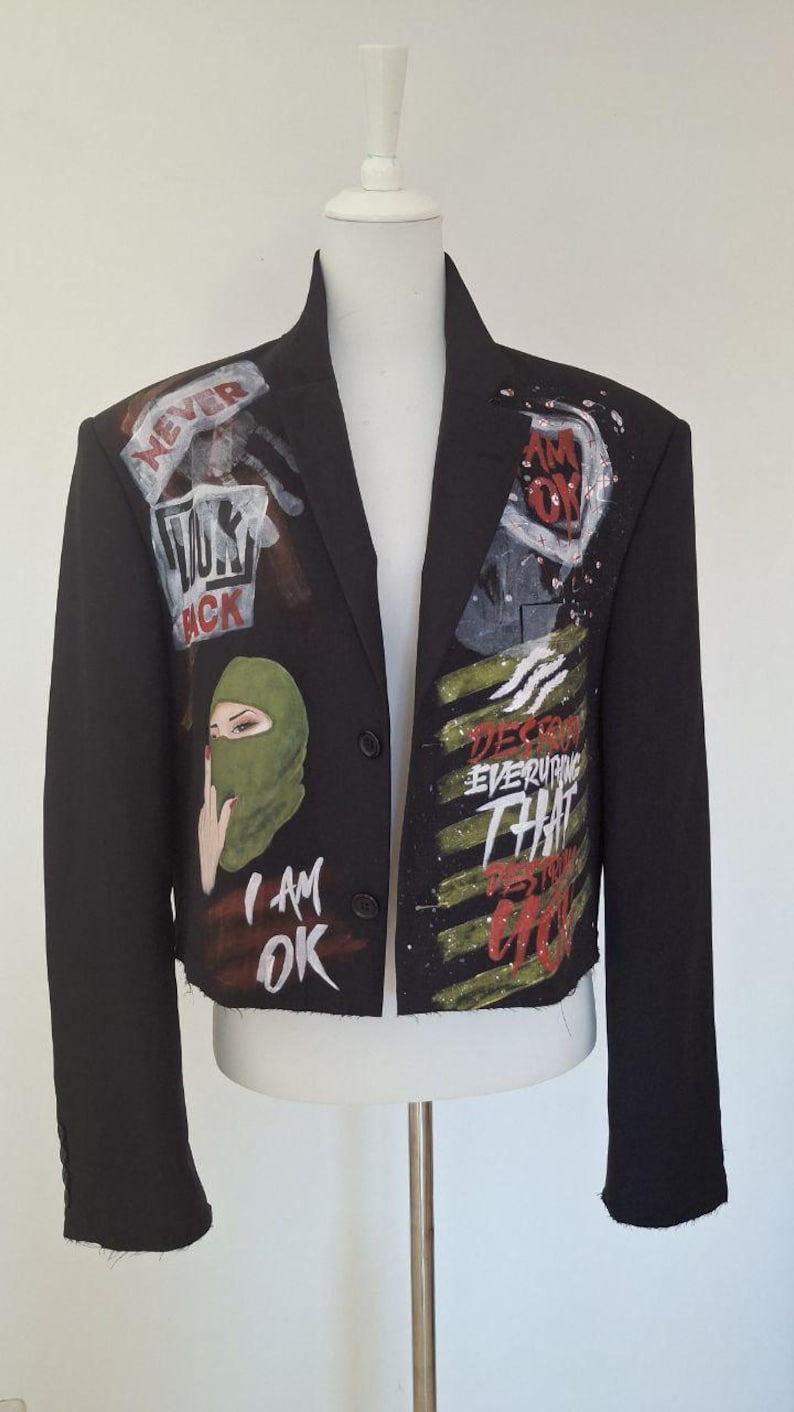 K&ouml;nnte beinhalten: Schwarzer Blazer mit handgemalten Kunstwerken. Die Jacke zeigt eine gr&uuml;ne Sturmhaube, die Worte "I AM OK" und andere Phrasen. Das Kunstwerk ist in Rot, Wei&szlig; und Gr&uuml;n gehalten. Der Blazer wird auf einer wei&szlig;en Schaufensterpuppe pr&auml;sentiert.