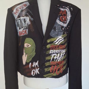 K&ouml;nnte beinhalten: Schwarzer Blazer mit bemalten Kunstwerken. Das Kunstwerk enth&auml;lt die Worte &bdquo;NEVER LOOK BACK&ldquo;, &bdquo;I AM OK&ldquo; und &bdquo;DESTROY EVERYTHING THAT DESTROYS YOU&ldquo;. Auf dem Blazer ist auch eine gr&uuml;ne Sturmhaube abgebildet.