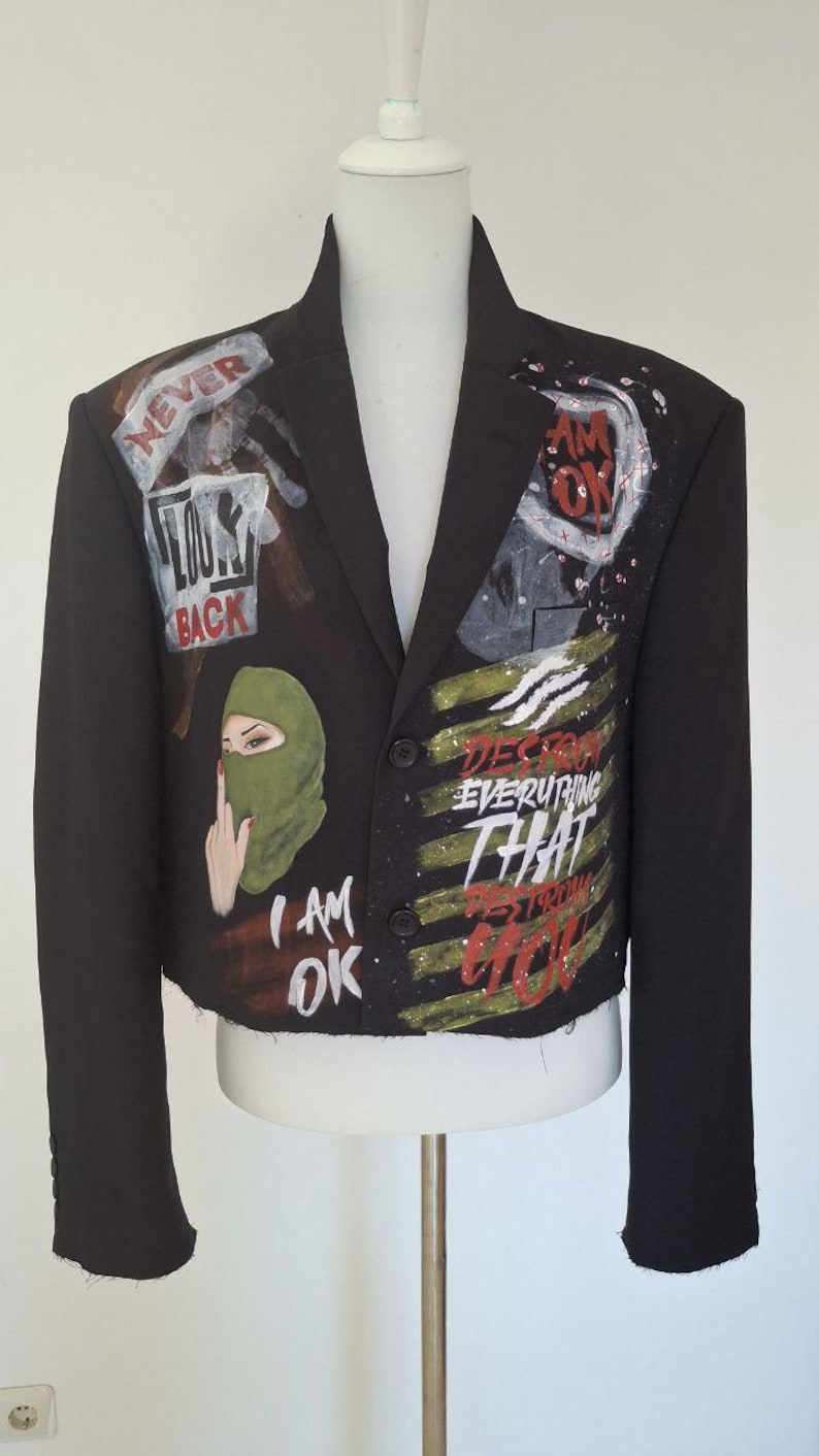 K&ouml;nnte beinhalten: Schwarzer Blazer mit bemalten Kunstwerken. Das Kunstwerk enth&auml;lt die Worte &bdquo;NEVER LOOK BACK&ldquo;, &bdquo;I AM OK&ldquo; und &bdquo;DESTROY EVERYTHING THAT DESTROYS YOU&ldquo;. Auf dem Blazer ist auch eine gr&uuml;ne Sturmhaube abgebildet.