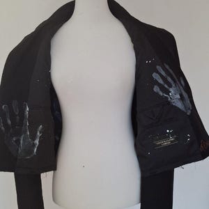 K&ouml;nnte beinhalten: Schwarzer Blazer mit wei&szlig;en Handabdruck-Designs auf dem Innenfutter. Der Blazer wird auf einer wei&szlig;en Schaufensterpuppe pr&auml;sentiert. Das Innenfutter hat auch wei&szlig;e Farbspritzer. Der Blazer hat einen klassischen Schnitt.