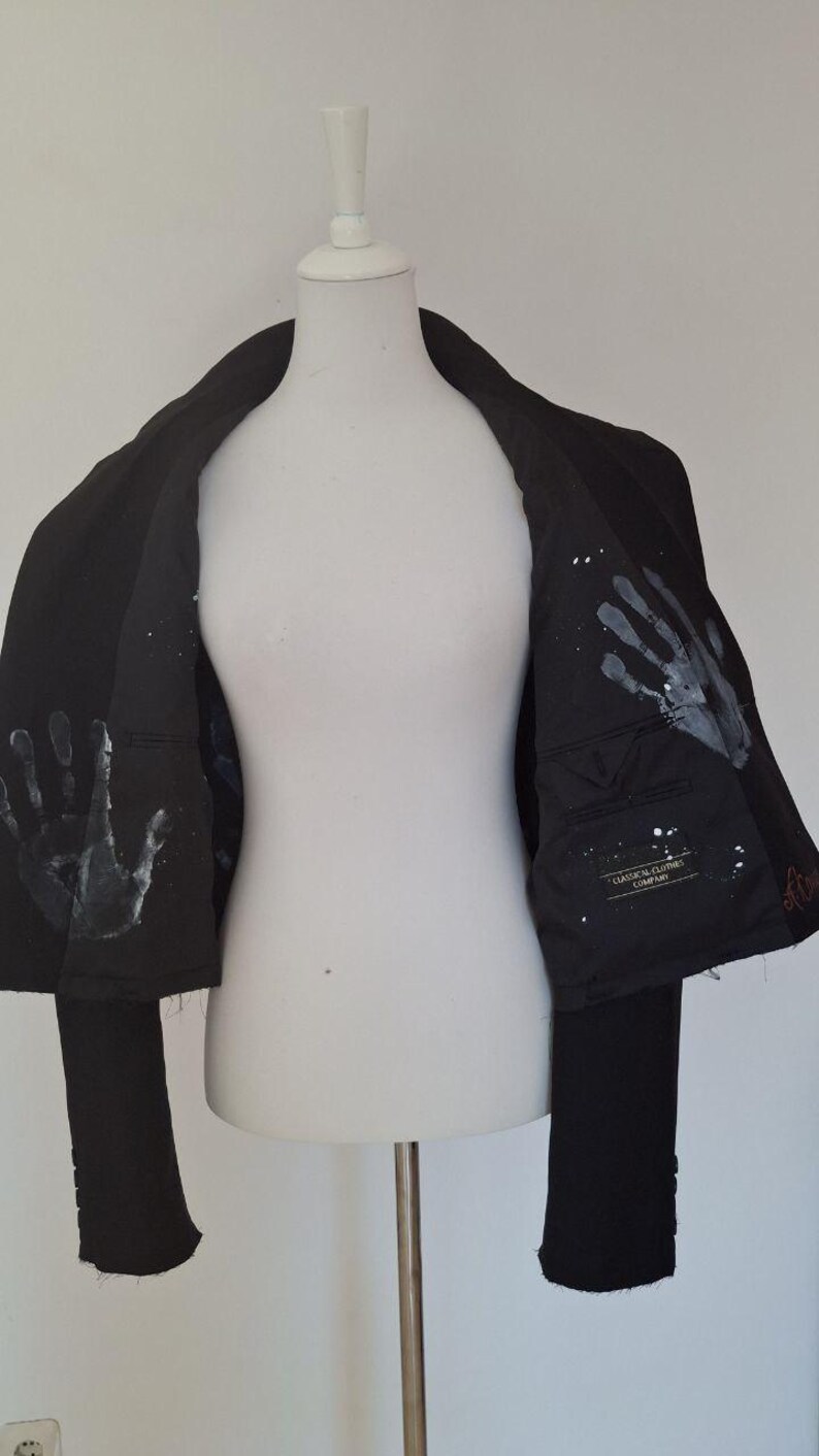 K&ouml;nnte beinhalten: Schwarzer Blazer mit wei&szlig;en Handabdruck-Designs auf dem Innenfutter. Der Blazer wird auf einer wei&szlig;en Schaufensterpuppe pr&auml;sentiert. Das Innenfutter hat auch wei&szlig;e Farbspritzer. Der Blazer hat einen klassischen Schnitt.