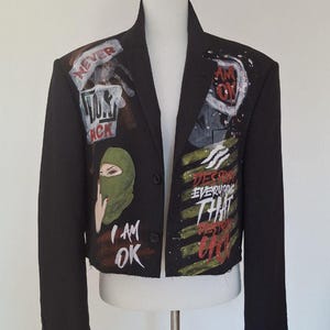K&ouml;nnte beinhalten: Schwarzer Blazer mit bemalten Kunstwerken. Die Jacke zeigt eine gr&uuml;ne Sturmhaube, die Worte "I AM OK" sowie weitere Texte und Grafiken in Wei&szlig;, Rot und Schwarz. Der Blazer wird auf einer wei&szlig;en Schaufensterpuppe pr&auml;sentiert.