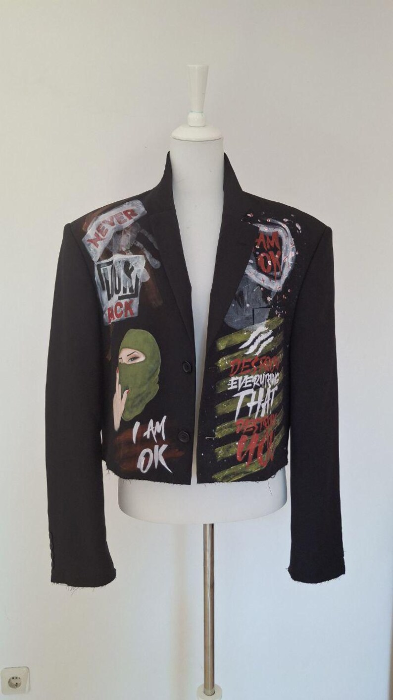 K&ouml;nnte beinhalten: Schwarzer Blazer mit bemalten Kunstwerken. Die Jacke zeigt eine gr&uuml;ne Sturmhaube, die Worte "I AM OK" sowie weitere Texte und Grafiken in Wei&szlig;, Rot und Schwarz. Der Blazer wird auf einer wei&szlig;en Schaufensterpuppe pr&auml;sentiert.