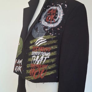K&ouml;nnte beinhalten: Schwarzer Blazer mit bemalten Kunstwerken. Das Kunstwerk enth&auml;lt die Worte "I AM OK" und "DESTROY EVERYTHING THAT STRONGER THAN YOU" in Rot, Wei&szlig; und Gr&uuml;n. Der Blazer wird auf einer Schaufensterpuppe pr&auml;sentiert.