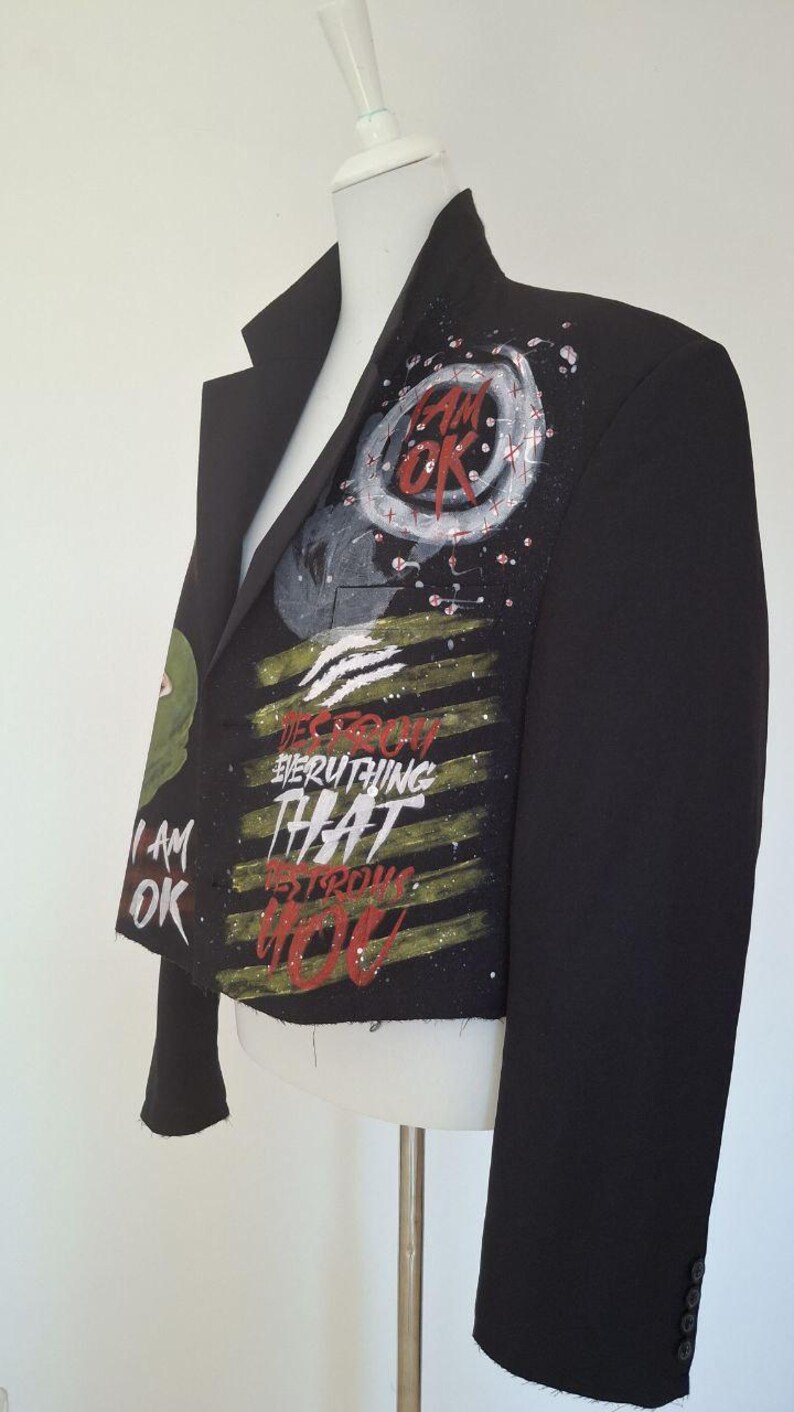 K&ouml;nnte beinhalten: Schwarzer Blazer mit bemalten Kunstwerken. Das Kunstwerk enth&auml;lt die Worte "I AM OK" und "DESTROY EVERYTHING THAT STRONGER THAN YOU" in Rot, Wei&szlig; und Gr&uuml;n. Der Blazer wird auf einer Schaufensterpuppe pr&auml;sentiert.