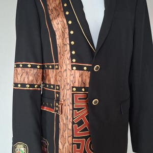 Könnte beinhalten: Schwarzer Blazer mit einem einzigartigen Holzmaserungsmuster und goldenen Akzenten. Der Blazer hat goldene Knöpfe und dekorative Elemente, mit einem Wappen auf dem Ärmel. Ein stilvolles und auffälliges Kleidungsstück.