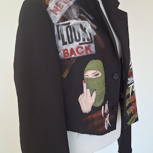K&ouml;nnte beinhalten: Schwarzer Blazer mit bemalten Kunstwerken. Das Kunstwerk enth&auml;lt die Worte "NEVR LOOK BACK" und eine Grafik einer Person mit gr&uuml;ner Sturmhaube und erhobenem Mittelfinger. Der Blazer hat einen klassischen Kragen und einen Knopfverschluss.