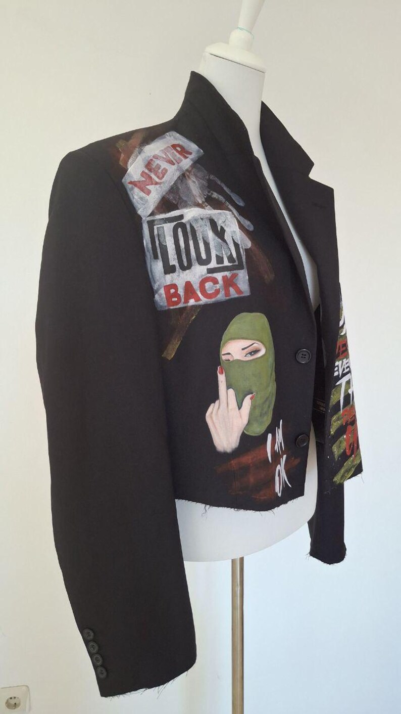 K&ouml;nnte beinhalten: Schwarzer Blazer mit bemalten Kunstwerken. Das Kunstwerk enth&auml;lt die Worte "NEVR LOOK BACK" und eine Grafik einer Person mit gr&uuml;ner Sturmhaube und erhobenem Mittelfinger. Der Blazer hat einen klassischen Kragen und einen Knopfverschluss.