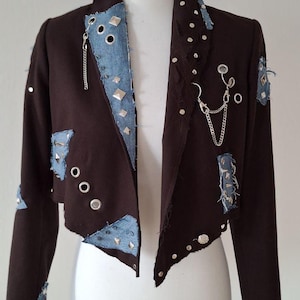 Könnte beinhalten: Eine braune, kurze Jacke mit Jeans-Patches und silbernen Nieten. Die Jacke hat einen Kragen und lange Ärmel. Die Jeans-Patches sind ausgefranst und haben einen Used-Look.