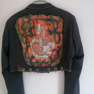 Puede incluir: Una chaqueta gris recortada con una pintura abstracta colorida de una cara en la espalda. La chaqueta tiene una fila de anillos plateados a lo largo del borde inferior.