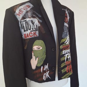 K&ouml;nnte beinhalten: Schwarzer Blazer mit bemalten Kunstwerken. Das Kunstwerk enth&auml;lt die Worte &bdquo;NEVER LOOK BACK&ldquo; und &bdquo;I AM OK&ldquo; sowie ein Bild einer Person mit einer gr&uuml;nen Sturmhaube. Der Blazer wird auf einer wei&szlig;en Schaufensterpuppe pr&auml;sentiert.