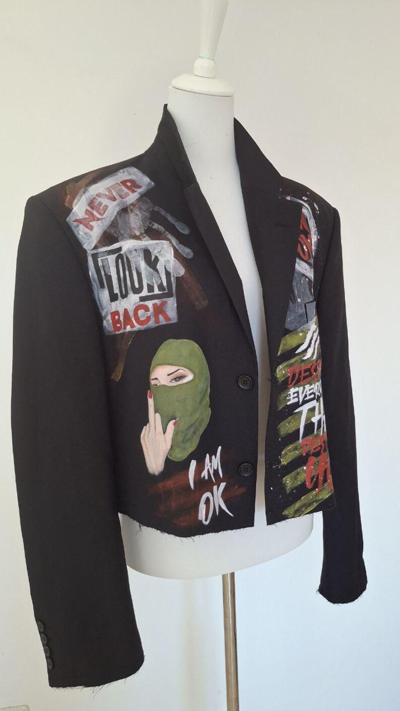 K&ouml;nnte beinhalten: Schwarzer Blazer mit bemalten Kunstwerken. Das Kunstwerk enth&auml;lt die Worte &bdquo;NEVER LOOK BACK&ldquo; und &bdquo;I AM OK&ldquo; sowie ein Bild einer Person mit einer gr&uuml;nen Sturmhaube. Der Blazer wird auf einer wei&szlig;en Schaufensterpuppe pr&auml;sentiert.