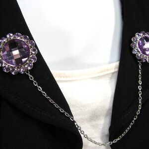 May include: Two decorative brooches linked by a chain. Each brooch has a large, faceted purple gemstone encircled by smaller purple stones. The brooches are fastened to a black jacket.