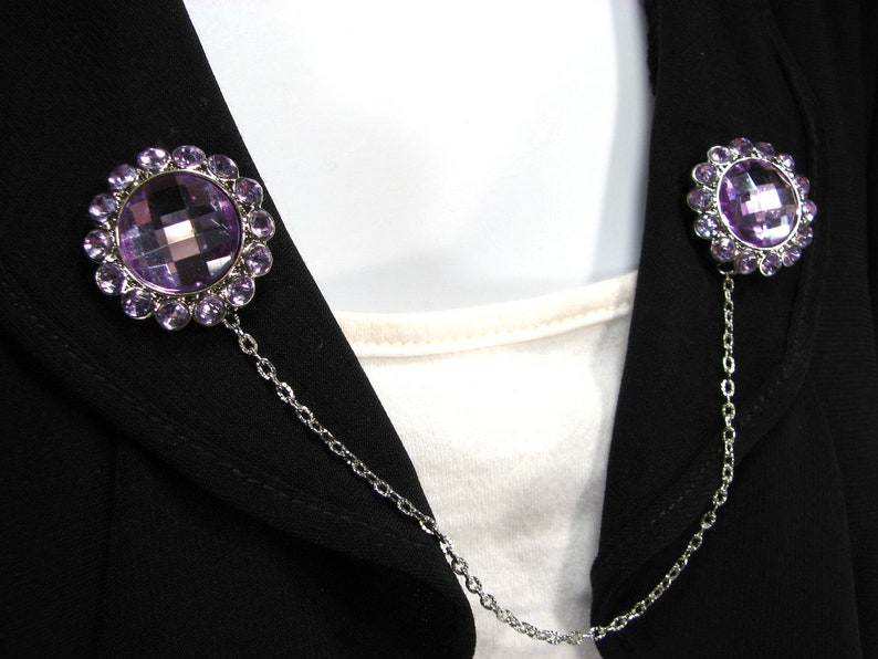 May include: Two decorative brooches linked by a chain. Each brooch has a large, faceted purple gemstone encircled by smaller purple stones. The brooches are fastened to a black jacket.