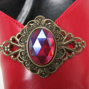 May include: A red leather shoe with a gold-toned metal buckle featuring a large, faceted, red gemstone.