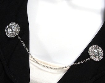 May include: A silver brooch set featuring two circular, clear crystal clusters linked by a fine chain. The brooches are fastened to a black jacket, providing a touch of sparkle and elegance to the outfit.