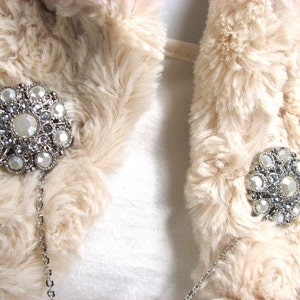 May include: A cream-colored faux fur jacket with two silver chains and large rhinestone brooches. The brooches are attached to the chains and are positioned on the front of the jacket.