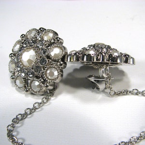 May include: A pair of silver tone brooches with a chain connecting them. Each brooch is round and features a cluster of white and clear rhinestones.