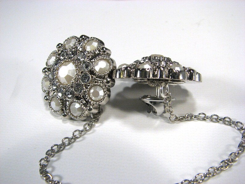 May include: A pair of silver tone brooches with a chain connecting them. Each brooch is round and features a cluster of white and clear rhinestones.