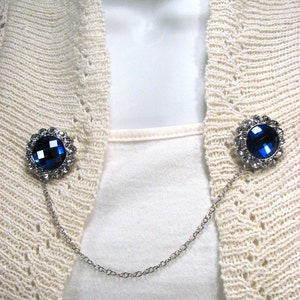 May include: A silver cardigan clip featuring two circular, dark blue jewel-like adornments. Each jewel is encircled by smaller clear crystals. A fine silver chain links the two embellishments, intended for securing a cardigan or sweater.