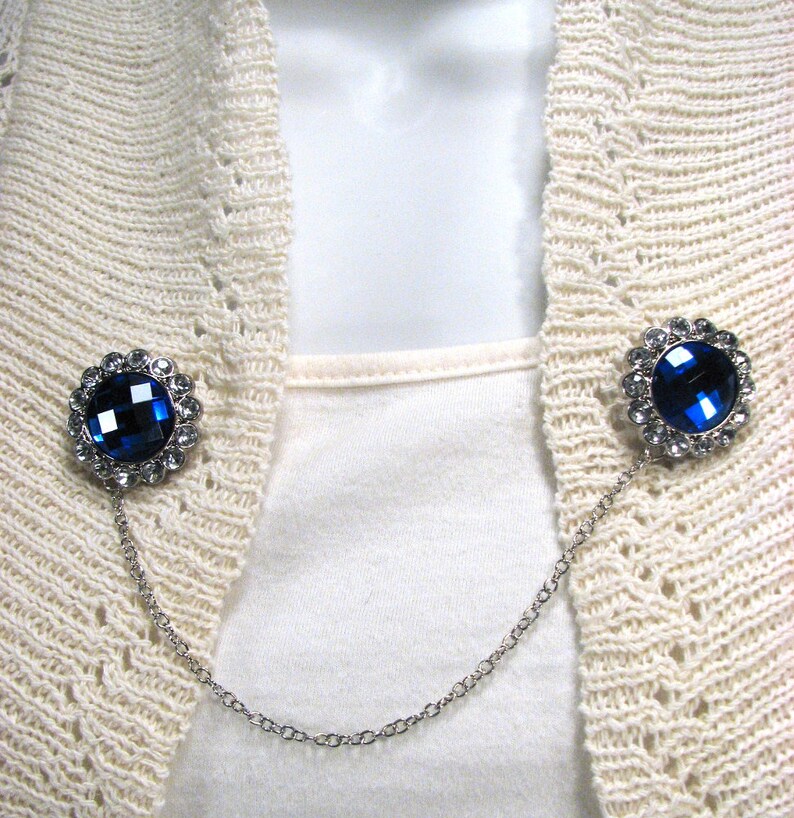 May include: A silver cardigan clip featuring two circular, dark blue jewel-like adornments. Each jewel is encircled by smaller clear crystals. A fine silver chain links the two embellishments, intended for securing a cardigan or sweater.
