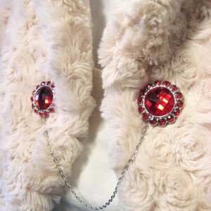 May include: A cream-coloured faux fur wrap featuring two red jewelled brooches linked by a silver chain. Each brooch has a large, faceted red centre stone encircled by smaller red gems.