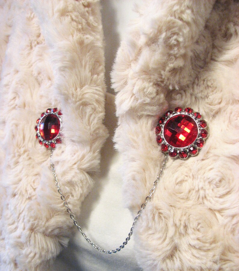 May include: A cream-coloured faux fur wrap featuring two red jewelled brooches linked by a silver chain. Each brooch has a large, faceted red centre stone encircled by smaller red gems.