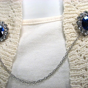 May include: A silver-toned sweater clip featuring two round, blue gemstone embellishments. Each gem is encircled by smaller, clear stones. A fine chain links the two decorative elements, designed to fasten a cardigan or sweater.