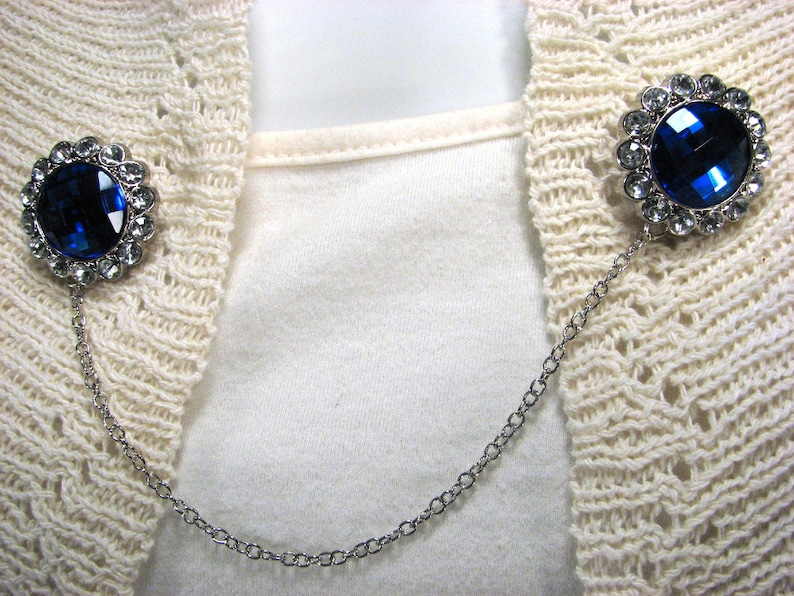 May include: A silver-toned sweater clip featuring two round, blue gemstone embellishments. Each gem is encircled by smaller, clear stones. A fine chain links the two decorative elements, designed to fasten a cardigan or sweater.