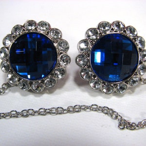 May include: Two silver cufflinks with large, faceted, deep blue central stones, encircled by clear, round crystals. Each cufflink is linked by a fine silver chain, offering a classic and elegant accessory, suitable for formal wear.