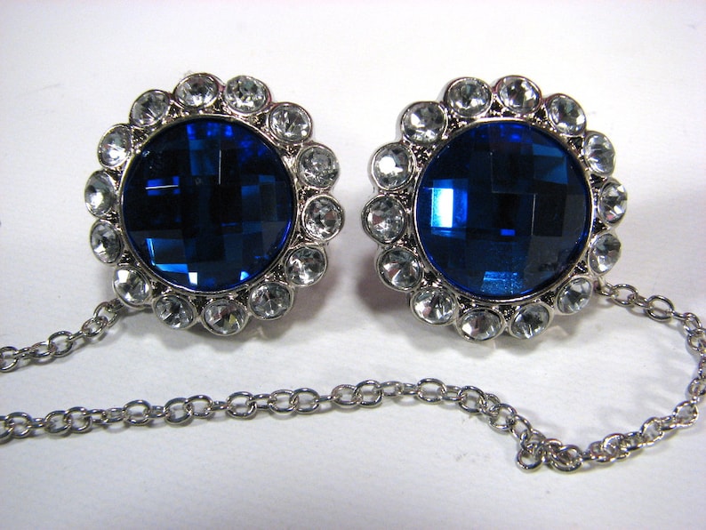 May include: Two silver cufflinks with large, faceted, deep blue central stones, encircled by clear, round crystals. Each cufflink is linked by a fine silver chain, offering a classic and elegant accessory, suitable for formal wear.