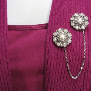 May include: Two silver-toned brooches with a chain connecting them. Each brooch features a circular design with white faux pearls and clear rhinestones.